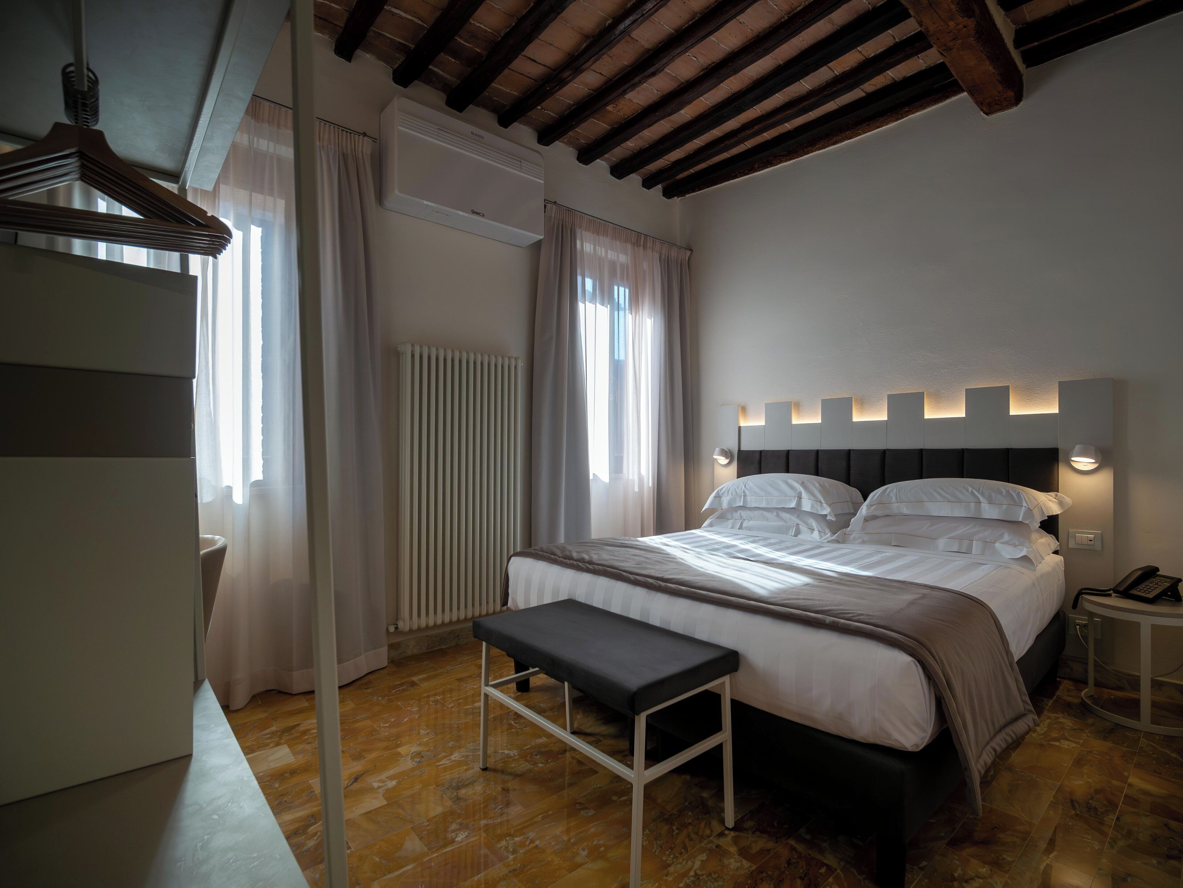 A bedroom with a bed, bedside tables, a bench, a radiator, and a window with curtains.