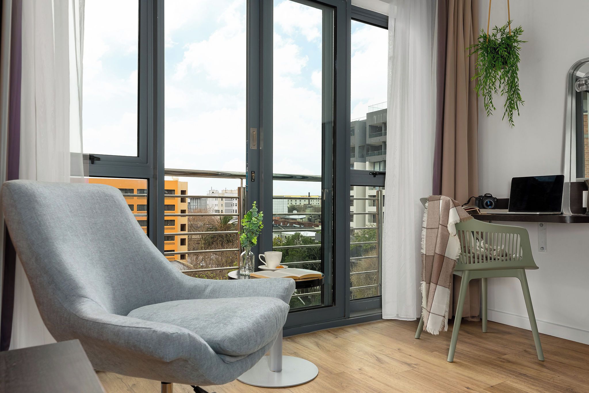 A modern living room with a grey armchair, a small table with a cup and book, a laptop on a desk, and a view of buildings through large glass doors.