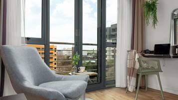A modern living room with a grey armchair, a small table with a cup and book, a laptop on a desk, and a view of buildings through large glass doors.