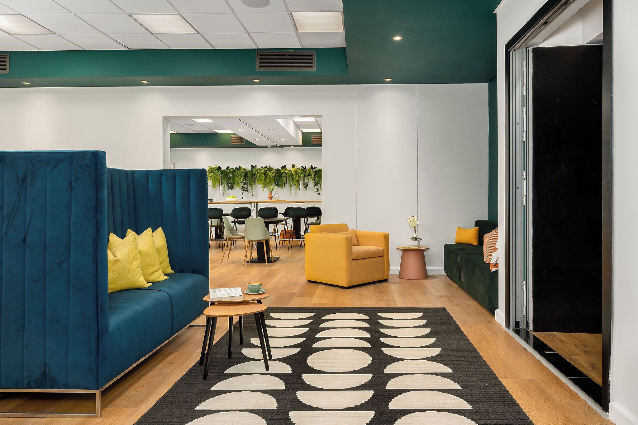 Eine moderne Lobby mit einem blauen Sofa, gelben Kissen, einem gelben Sessel, einem schwarz-weißen Teppich und einer grünen Wand mit Pflanzen.