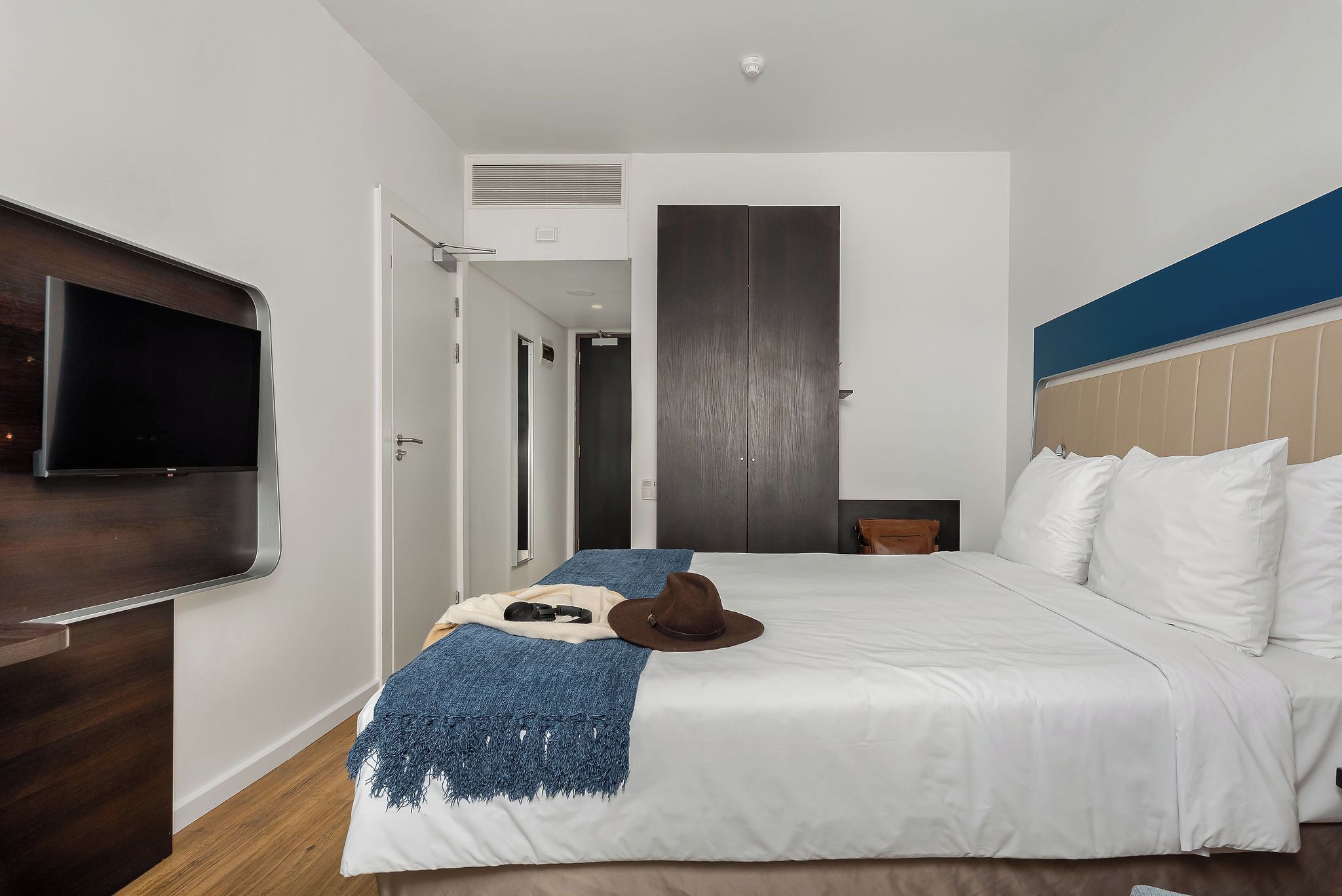 A hotel room with a bed, a television mounted on the wall, a wardrobe, and a door leading to another room.
