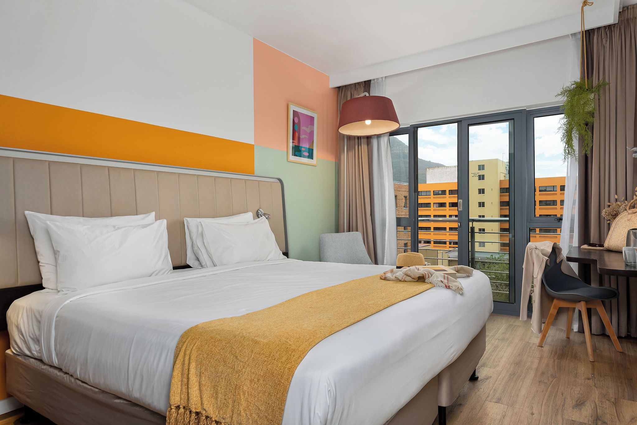 Een hotelkamer met een bed, een bureau, een stoel, een raam met uitzicht op gebouwen en een hangende plant.