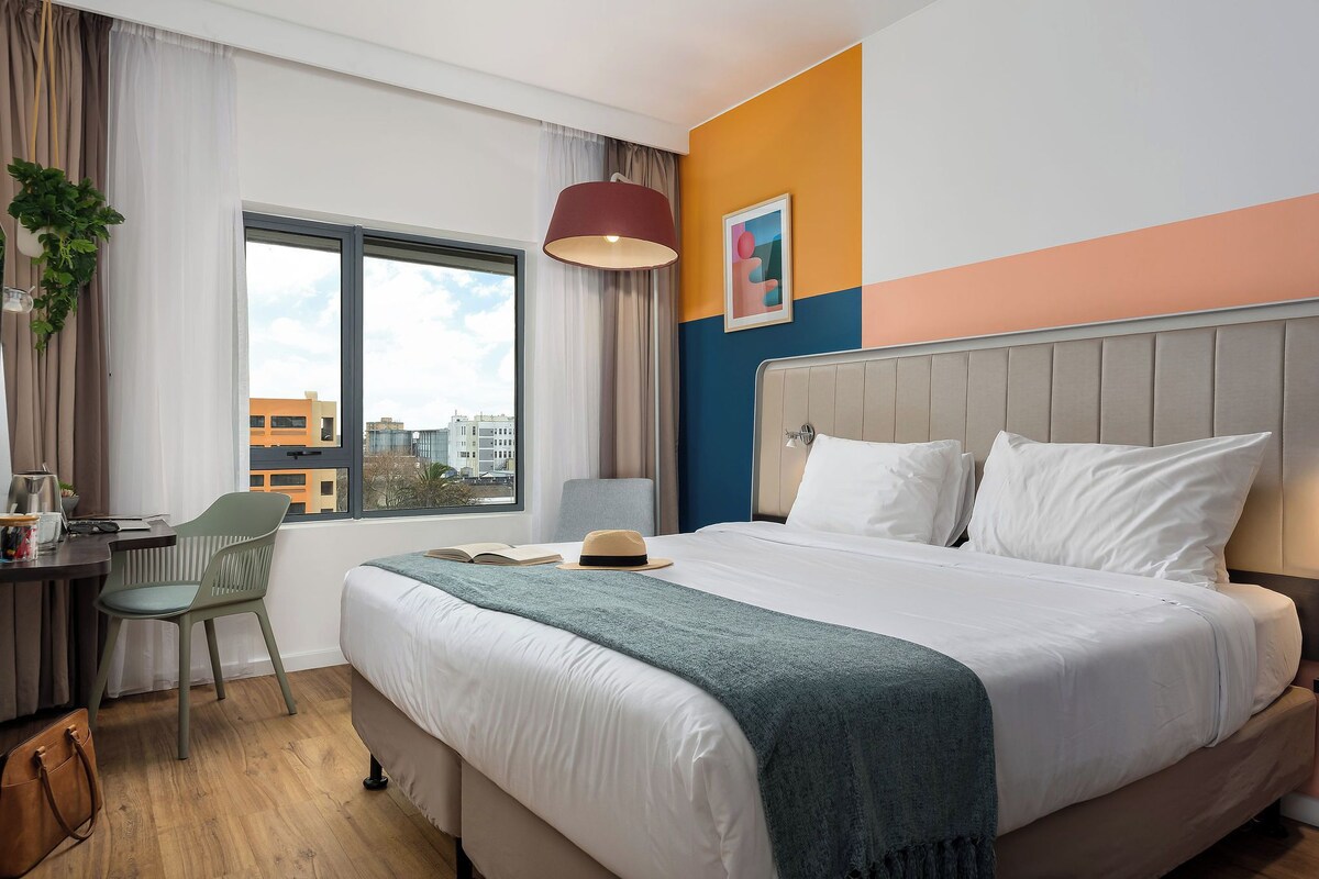 A hotel room with a bed, a desk, a chair, a window with a view of buildings, and a painting on the wall.
