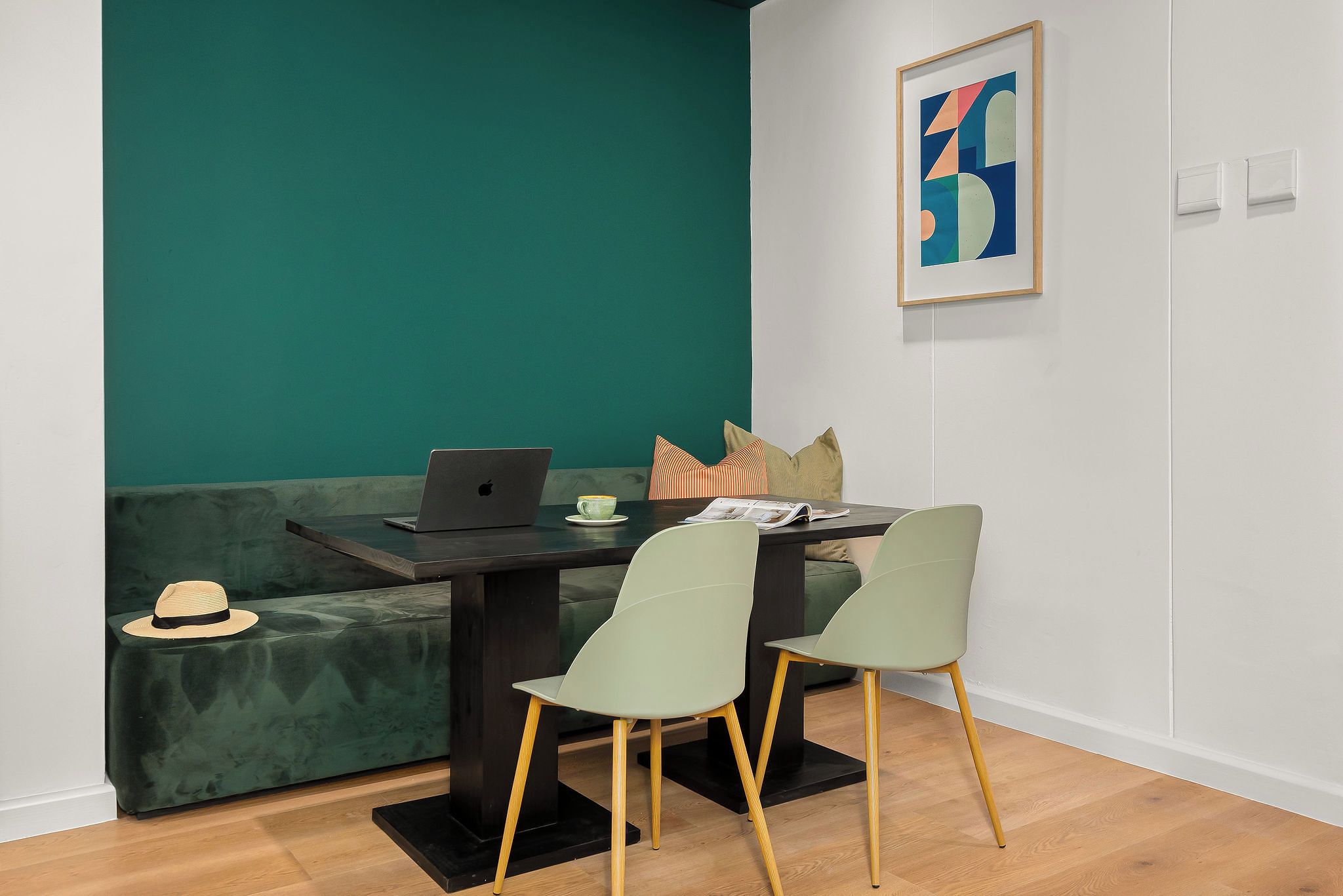 A modern room with a teal accent wall, a black table, two chairs, a laptop, a hat, and a framed abstract artwork.