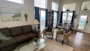 Un salon spacieux avec une grande table basse en verre, un canapé et deux fauteuils. On y trouve des ventilateurs de plafond, de grandes fenêtres avec des rideaux et une vue sur l’extérieur.