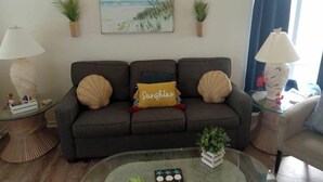 Un salon avec un canapé gris foncé, deux tables d’appoint avec des lampes, une table basse en verre ornée d’une plante et d’un plateau de biscuits, et un tableau sur le mur représentant un paysage marin.