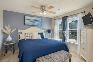 Un dormitorio con cama, ventilador de techo, ventana con cortinas, televisor y mesita de noche con lámpara.