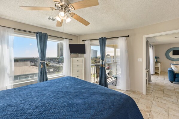 A bedroom with a large bed, a ceiling fan, a TV, and a balcony with a view of the city.