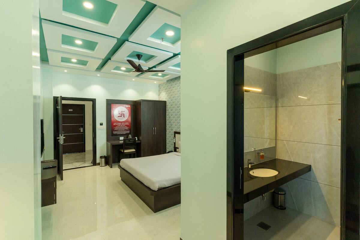 A modern hotel room with a bed, a desk, a wardrobe, and a bathroom with a sink and mirror.