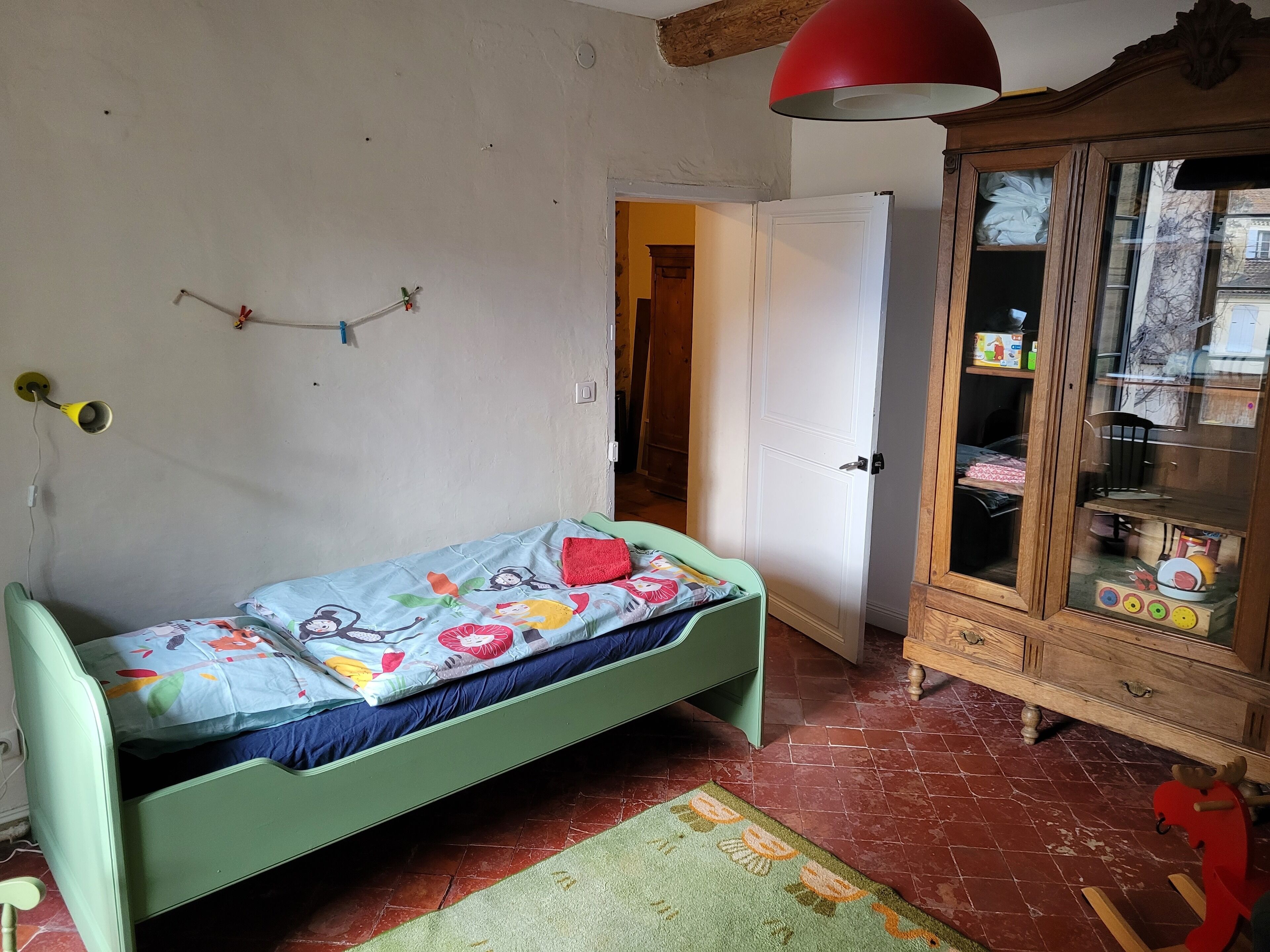 A small, single-bed room with a green bed, a wooden cabinet with glass doors, and a red hanging lamp.