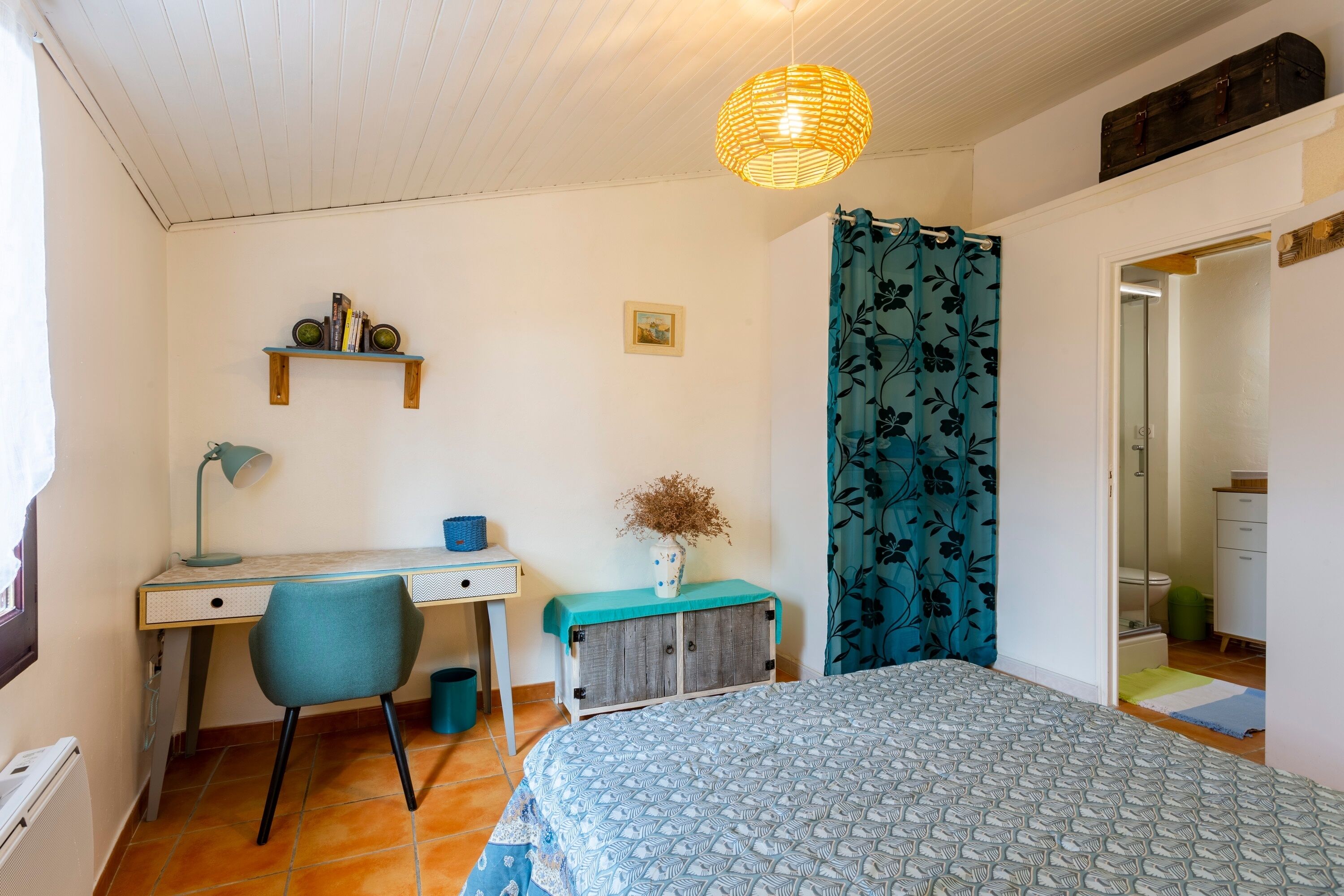 Ein Schlafzimmer mit einem Bett, einem Schreibtisch, einem Stuhl, einem Bücherregal, einer Vase mit Trockenblumen und einer Pendelleuchte.
