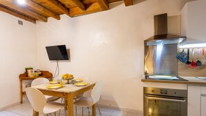 A kitchen with a wooden table, white chairs, a built-in oven, and a TV mounted on the wall. - L'Observance - Au cœur du centre historique (Arles)