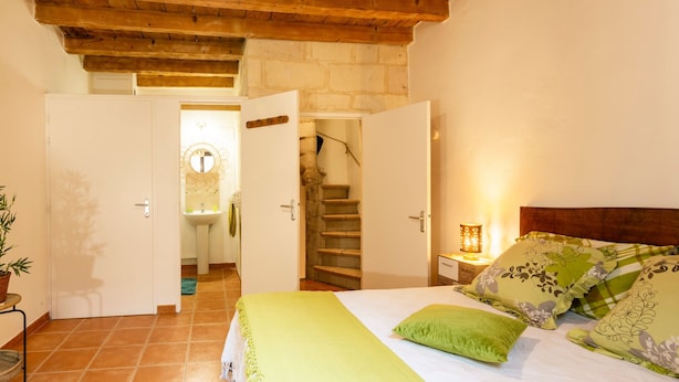 A bedroom with a bed, green pillows, a nightstand with a lamp, a wooden headboard, and a staircase leading to a bathroom. - L'Observance - Au cœur du centre historique (Arles)