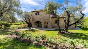A rustic stone building with arched doorways and a tiled roof, surrounded by lush greenery and flowering plants.