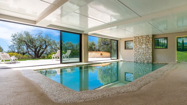A modern indoor swimming pool with clear glass walls, a stone wall, and a view of the outdoors.