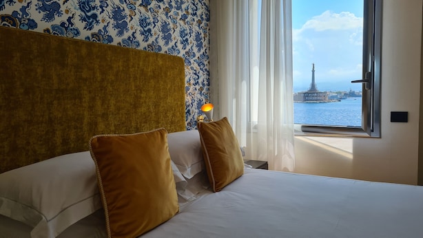 A hotel room with a large bed, two brown pillows, a headboard, and a window with a view of a distant cityscape and a tower. - Jolly Charme Suite (Messina)