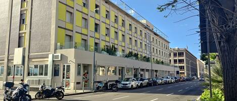 Un edificio moderno con fachada plana, ventanas grandes y un estacionamiento con coches y motocicletas.