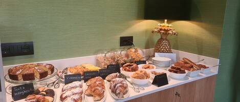 Buffet com diversos pastéis e sobremesas, incluindo tartes, croissants e muffins, expostos em um balcão com etiquetas.