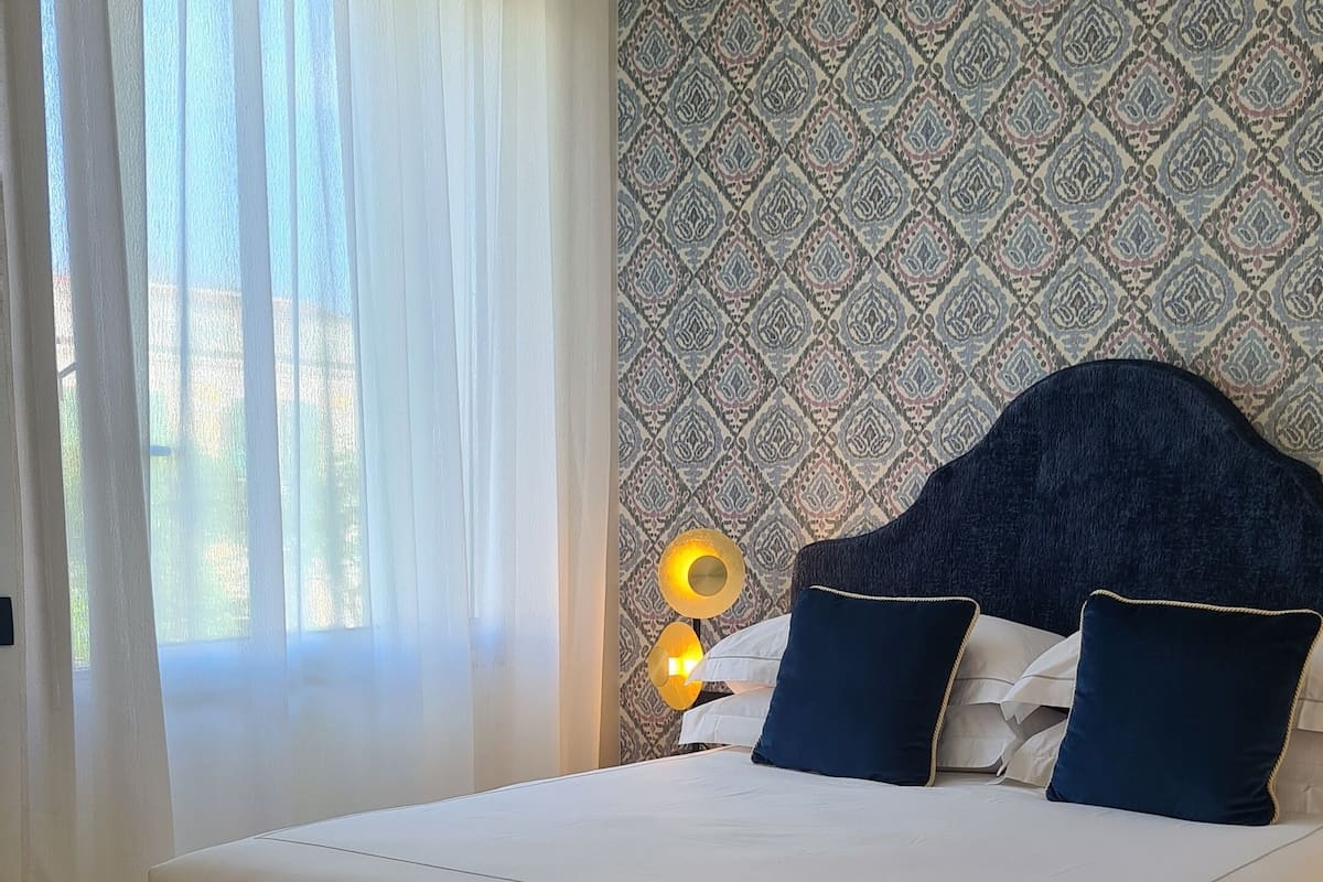 Ein Schlafzimmer mit einem großen Bett, einer Bank, einem gemusterten Wandteppich und einem Fenster mit Vorhängen.