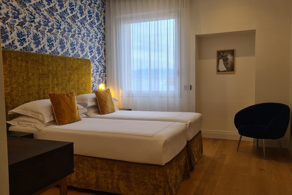 Ein Hotelzimmer mit zwei Betten, einem Schreibtisch, einem Stuhl und einem Fenster mit durchsichtigen Vorhängen.