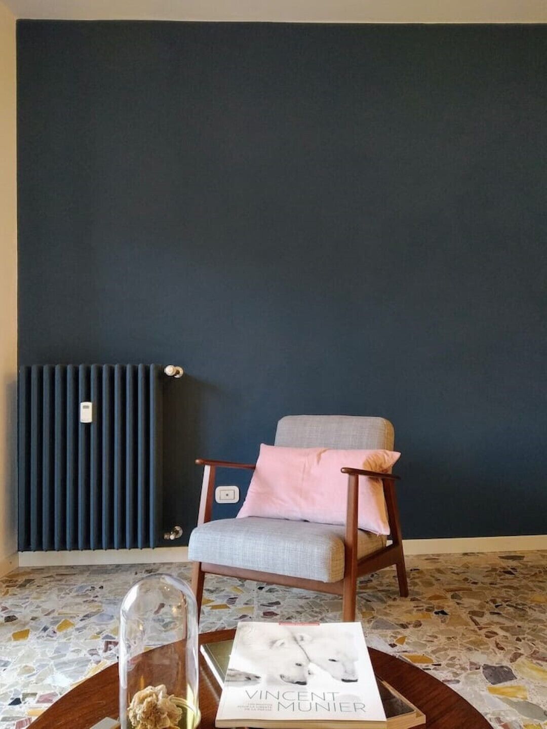 Una habitación con pared azul oscuro, un radiador, un sillón gris y un libro titulado 'Vincent Munier' sobre una mesa.