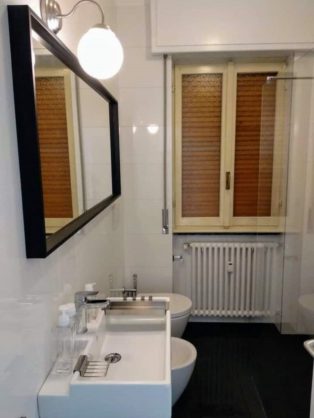 A modern bathroom with a white sink, mirror, and toilet. There is a window with wooden blinds and a radiator below.