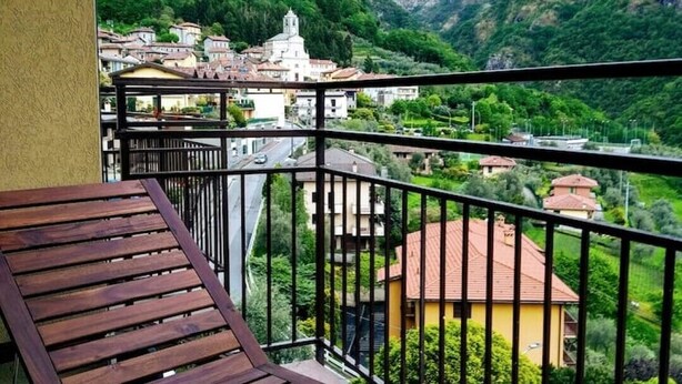 A balcony with a wooden floor and black railing, overlooking a town with buildings and green hills. - Renovated, amazing lake view (Perledo)