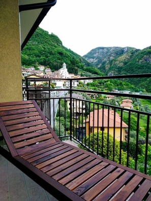 A balcony with a wooden floor and black railing, overlooking a town with buildings and green hills. - Renovated, amazing lake view (Perledo)