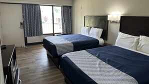 A hotel room with two beds, a desk, a chair, a window with curtains, and a wall-mounted lamp. - Oakwood Inn (Oakdale)