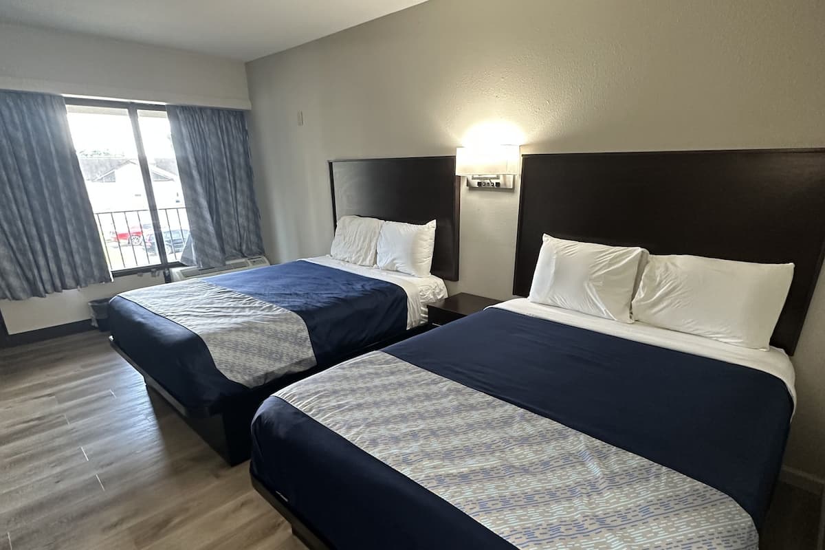 A hotel room with two beds, a large window with curtains, a wooden floor, and a wall-mounted light fixture.