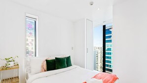 A hotel room with a bed, a window with a view of buildings, and a plant on a shelf.