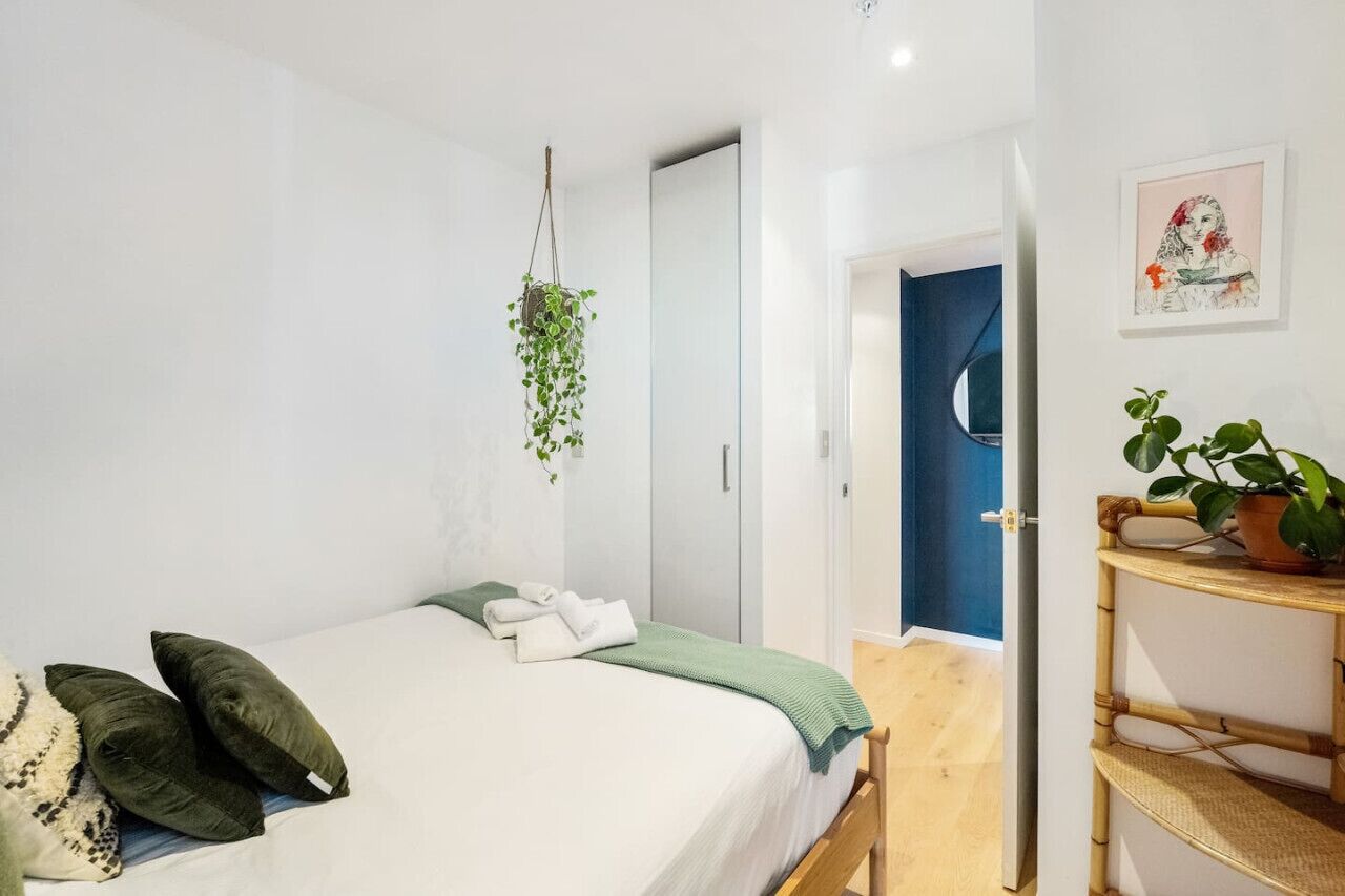 A bedroom with a bed, pillows, a hanging plant, a wardrobe, and a door leading to another room.