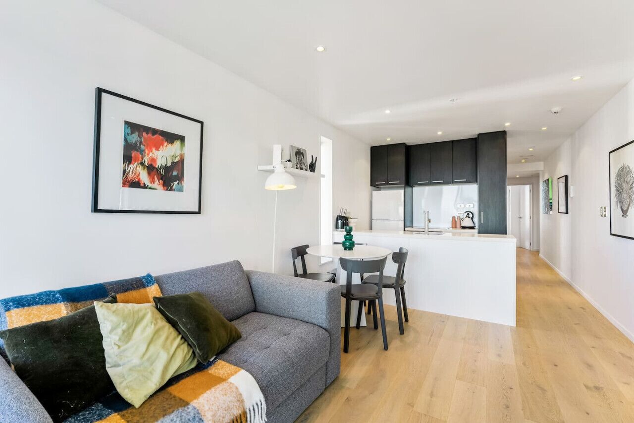 Un salon moderne avec un canapé gris, une table basse et un coin repas comprenant une table et des chaises blanches. On aperçoit, au fond, une cuisine avec des meubles sombres et un plan de travail blanc.