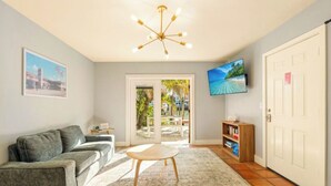 A living room with a grey sofa, a small wooden table, a TV mounted on the wall, and a door leading to an outdoor area.