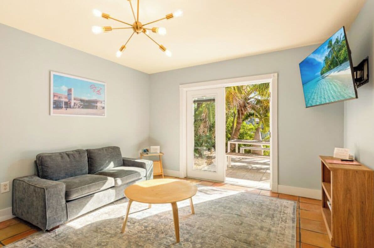 A living room with a grey sofa, a wooden coffee table, a TV mounted on the wall, and a sliding glass door leading to an outdoor area.
