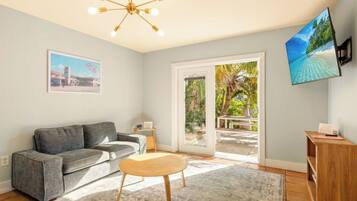 A living room with a grey sofa, a wooden coffee table, a TV mounted on the wall, and a sliding glass door leading to an outdoor area.