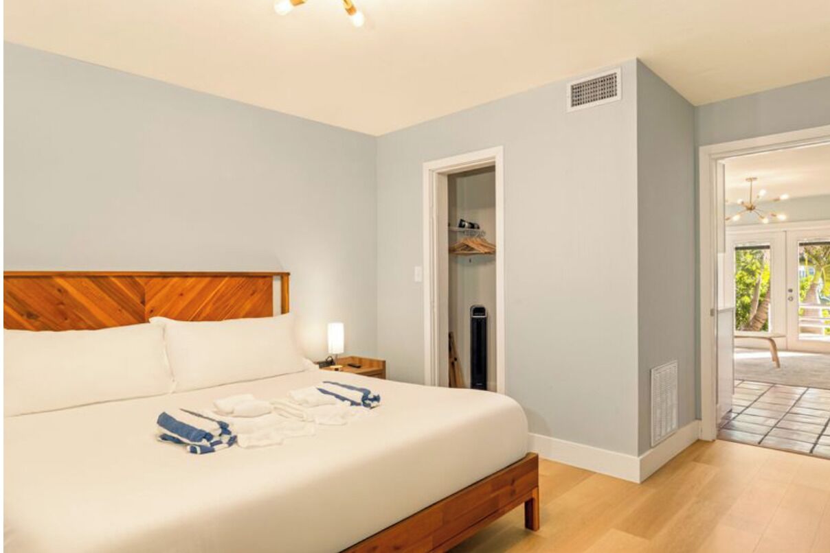 A bedroom with a wooden headboard, a bed with white linens, a nightstand, and a closet with hanging clothes.