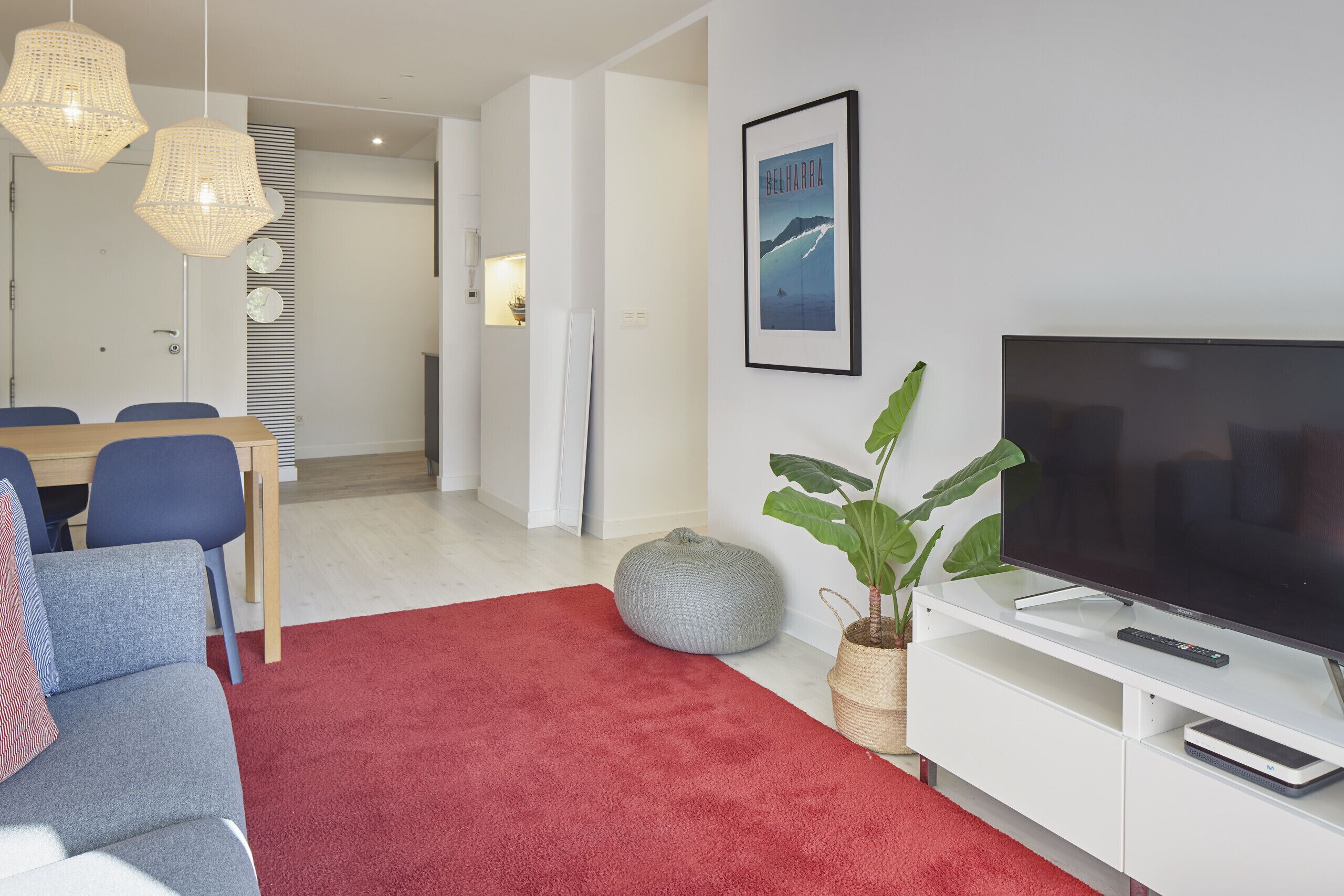 Un salon moderne avec une moquette rouge, un grand téléviseur à écran plat, un meuble de rangement blanc, un pouf gris et une grande plante verte.