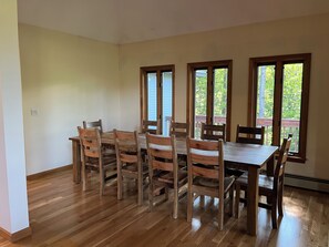 Een eetruimte met een houten tafel en stoelen, grote ramen met uitzicht op groen en een houten vloer.