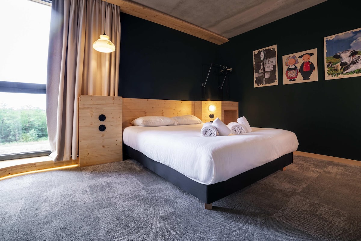 Habitación de hotel con cama, ventana con cortinas, lámpara de pared y una obra de arte.