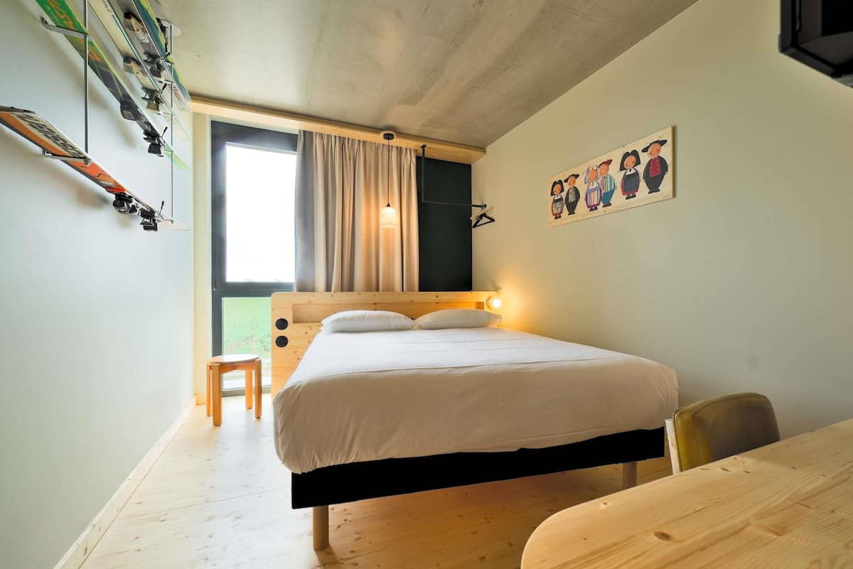 A bedroom with a bed, a wooden desk, a chair, a window with curtains, and a wall-mounted ski rack.