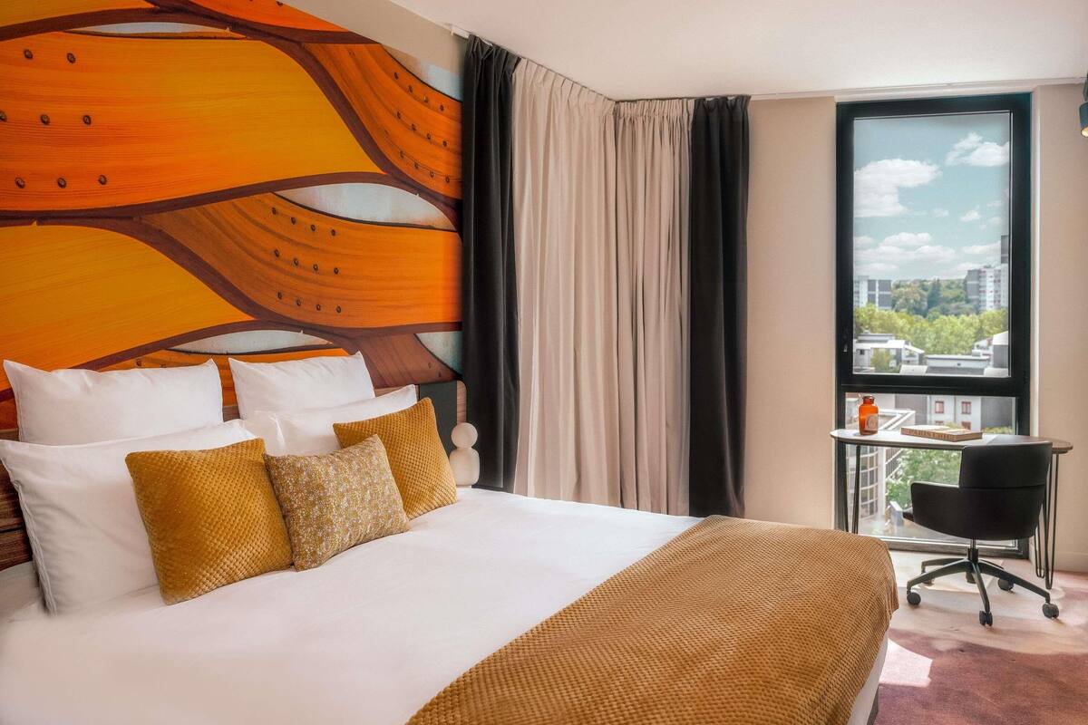A hotel room with a large bed, a view of the city, and a small table with a chair.