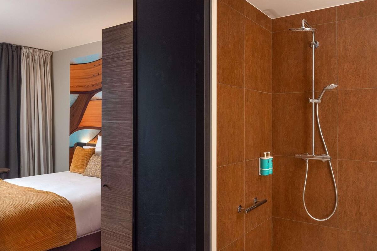 A hotel room with a bed, a wooden accent wall, a shower with a handheld showerhead, and a towel rack.
