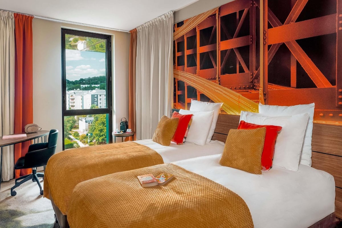 Habitación de hotel con dos camas, un escritorio y vistas a edificios a través de un ventanal grande.