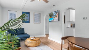 Ein modernes Wohnzimmer mit einem blauen Sofa, einem runden Holztisch und einem Esstisch mit Stühlen. An den Wänden befinden sich gerahmte Kunstwerke und ein Fernseher, der über dem Sofa angebracht ist.