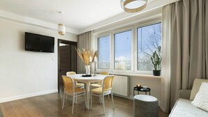 A modern living room with a dining area, large windows, a flat-screen TV, and a potted plant. - Elite Apartments Sunset (Gdansk)