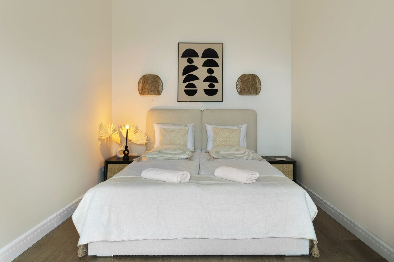 Una cama bien hecha con colcha blanca y dos almohadas, una lámpara de noche y una obra de arte abstracta colgada en la pared.