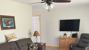 Ein Wohnzimmer mit grauem Sofa, zwei schwarzen Ledersesseln, einer Holzkabine, einem Fernseher, einem Deckenventilator und einem gerahmten Bild an der Wand.
