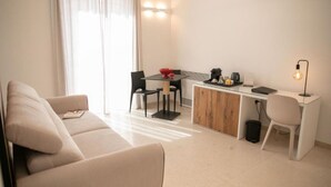 A modern living room with a sofa, a dining table with chairs, a small kitchenette, and a desk with a lamp. - KALEA Guest House (Cefalù)