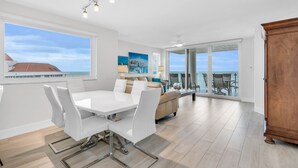 Een ruime eethoek met een witte tafel en stoelen, een bank met kussens, en uitzicht op de oceaan door grote ramen.